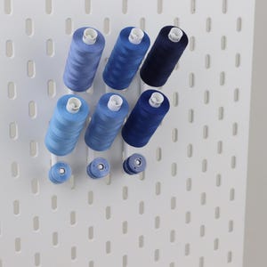 May include: A white pegboard wall organizer featuring three vertical rows of blue sewing thread spools in varying shades, ranging from light sky blue to deep navy blue, mounted on white pegs for storage and organization of craft supplies.