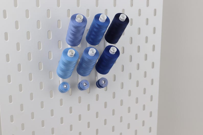 May include: A white pegboard wall organizer featuring three vertical rows of blue sewing thread spools in varying shades, ranging from light sky blue to deep navy blue, mounted on white pegs for storage and organization of craft supplies.