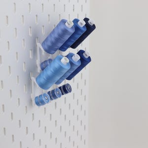 May include: A white pegboard wall organizer featuring several blue sewing thread spools of varying sizes arranged on white plastic pegs. The spools are organized in rows, displaying shades of light blue, medium blue, and dark navy blue thread.