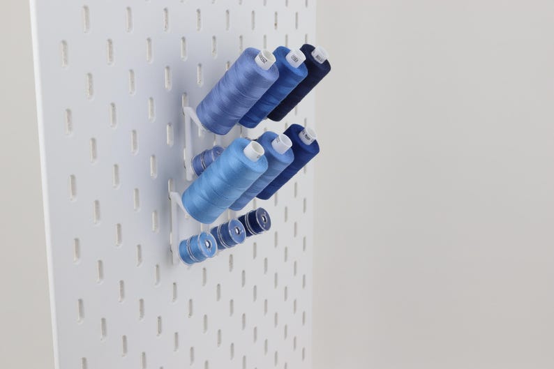 May include: A white pegboard wall organizer featuring several blue sewing thread spools of varying sizes arranged on white plastic pegs. The spools are organized in rows, displaying shades of light blue, medium blue, and dark navy blue thread.