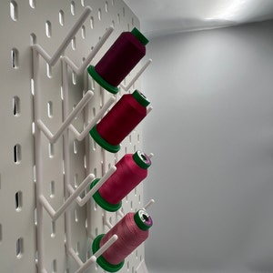 Pu&ograve; includere: Un pannello forato bianco con quattro bobine di filo appese. Le bobine sono rosa, rosse e viola. Le bobine sono appese a ganci sul pannello forato.