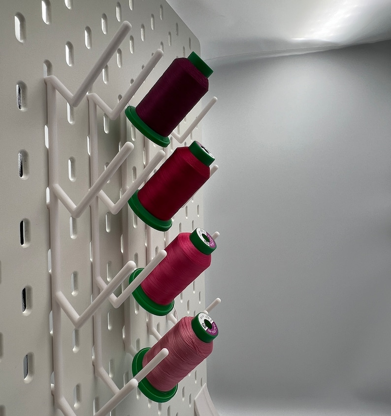Pu&ograve; includere: Un pannello forato bianco con quattro bobine di filo appese. Le bobine sono rosa, rosse e viola. Le bobine sono appese a ganci sul pannello forato.