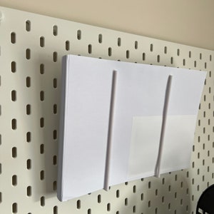 May include: A stack of white paper is held in place by two white plastic rods on a white pegboard wall.