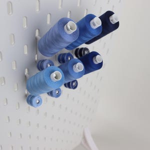 May include: A white pegboard panel featuring a collection of sewing thread spools in various shades of blue, ranging from light sky blue to deep navy. The spools are mounted on white pegs, organized in a cluster on the perforated surface.