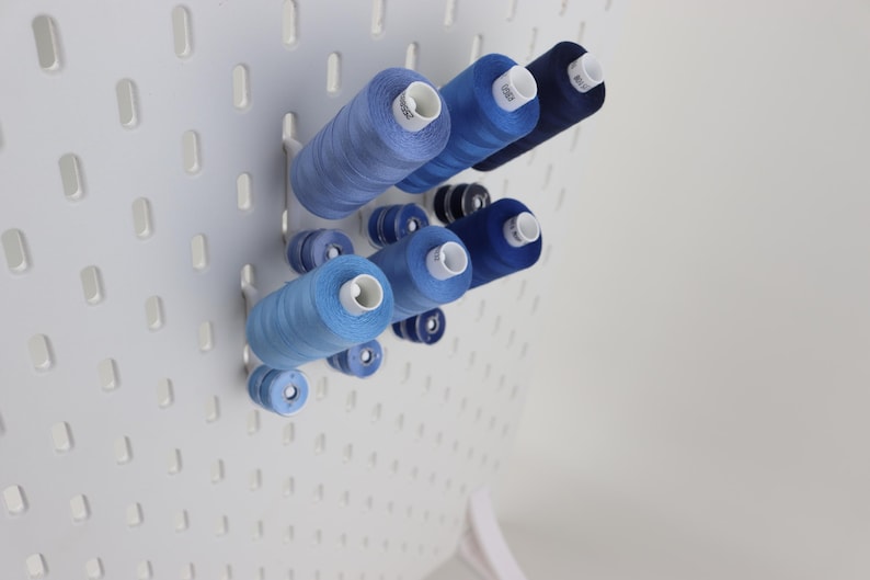 May include: A white pegboard panel featuring a collection of sewing thread spools in various shades of blue, ranging from light sky blue to deep navy. The spools are mounted on white pegs, organized in a cluster on the perforated surface.