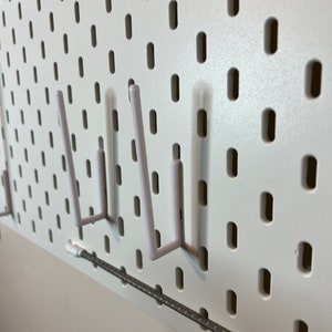 May include: White pegboard with three white plastic hooks. The hooks are in the shape of a J and are attached to the pegboard.