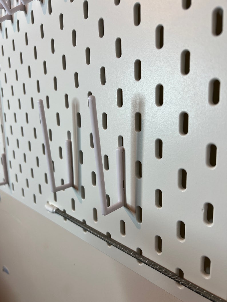 May include: White pegboard with three white plastic hooks. The hooks are in the shape of a J and are attached to the pegboard.