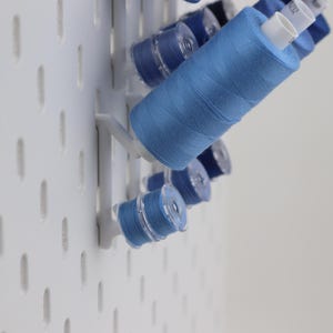 May include: A white pegboard wall organizer featuring several blue sewing thread spools and bobbins mounted on white plastic pegs. The spools are arranged in a vertical pattern, showcasing various shades of blue thread wound onto plastic cores.