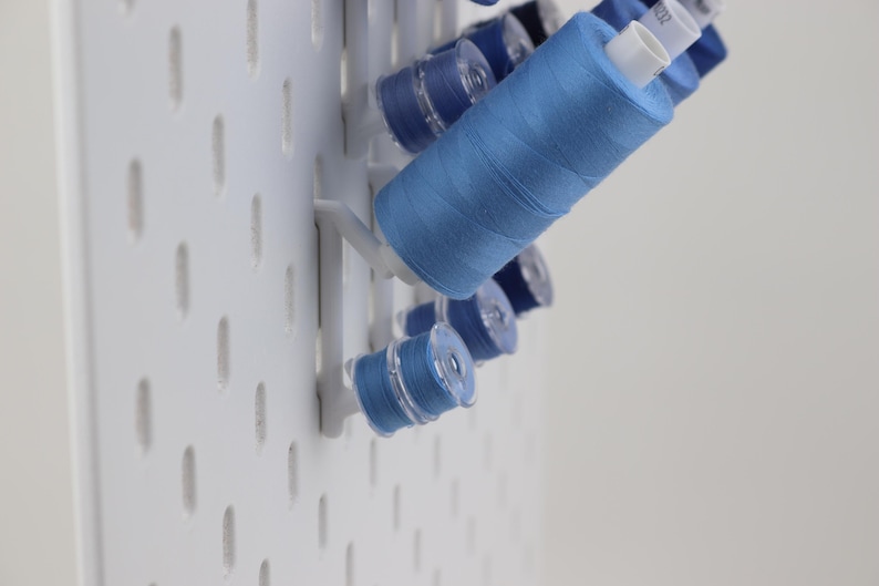 May include: A white pegboard wall organizer featuring several blue sewing thread spools and bobbins mounted on white plastic pegs. The spools are arranged in a vertical pattern, showcasing various shades of blue thread wound onto plastic cores.
