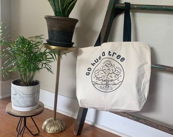 Könnte beinhalten: Eine weiße Canvas-Tragetasche mit schwarzen Riemen und dem Aufdruck "Go hug a tree" auf der Vorderseite. Die Tasche zeigt eine Schwarzweißillustration eines Baumes mit Wurzeln und Blättern. Die Tasche hängt an einer Holzleiter.