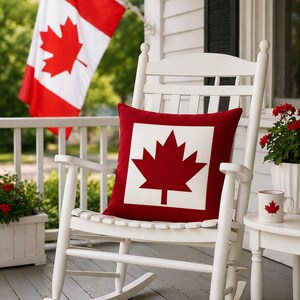 May include: A white wooden rocking chair with a red and white pillow featuring a maple leaf design. A Canadian flag waves in the background. A matching mug with a maple leaf sits on a small side table. Red flowers are in a white planter.