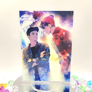 May include: A clear acrylic stand with a printed image of two men. The man on the left is wearing a plaid shirt and the man on the right is wearing a red hat and a red and white jacket. The background is a blurred image of a city skyline.