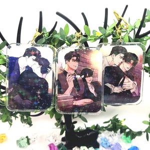 May include: Three clear acrylic keychains with images of two men in different poses. The keychains are hanging from a green leafy branch.