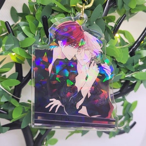 Op de afbeelding: Een heldere acryl sleutelhanger met een kleurrijke illustratie van twee anime-achtige personages die elkaar omarmen. De sleutelhanger heeft een goudkleurige hartvormige sluiting en een holografisch effect. De achtergrond bevat kunstgroen en een decoratieve boom.