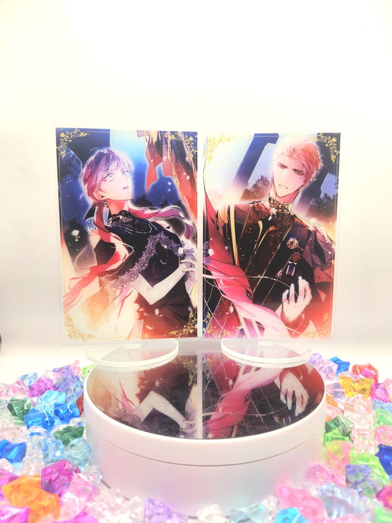 May include: Two anime-style character art prints, one with pink hair and the other with blonde hair, displayed on a white rotating stand. The stand is surrounded by colourful, transparent, decorative stones.