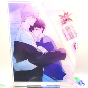 May include: A clear acrylic stand featuring a colorful illustration of two figures embracing. One figure has light purple hair and is wearing a white shirt, while the other has dark hair and is wearing a blue shirt. A small plant and a gift box are visible.
