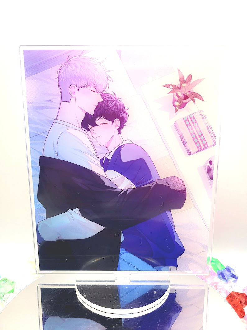 May include: A clear acrylic stand featuring a colorful illustration of two figures embracing. One figure has light purple hair and is wearing a white shirt, while the other has dark hair and is wearing a blue shirt. A small plant and a gift box are visible.