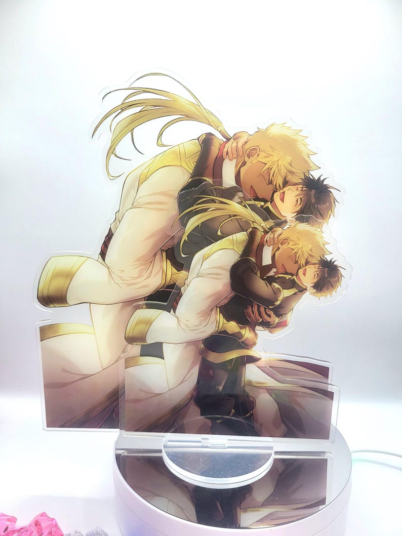 May include: A clear acrylic stand with a printed image of two anime characters embracing. The characters are both male and have blonde hair. The character in the front is wearing a black and gold outfit, while the character in the back is wearing a white and gold outfit.