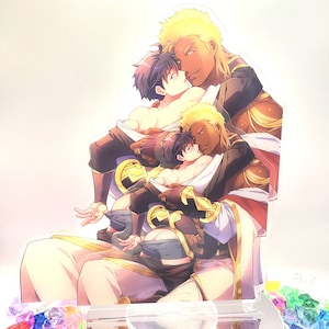 May include: A three-dimensional acrylic stand featuring a colorful illustration of three men embracing. The men are depicted with different hair colors and skin tones, wearing elaborate clothing with gold accents. The stand is designed to hold a phone or other small object.