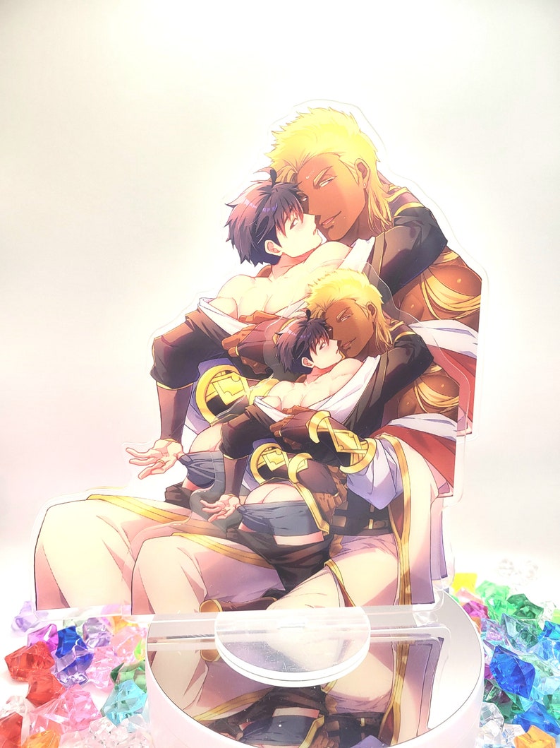 May include: A three-dimensional acrylic stand featuring a colorful illustration of three men embracing. The men are depicted with different hair colors and skin tones, wearing elaborate clothing with gold accents. The stand is designed to hold a phone or other small object.