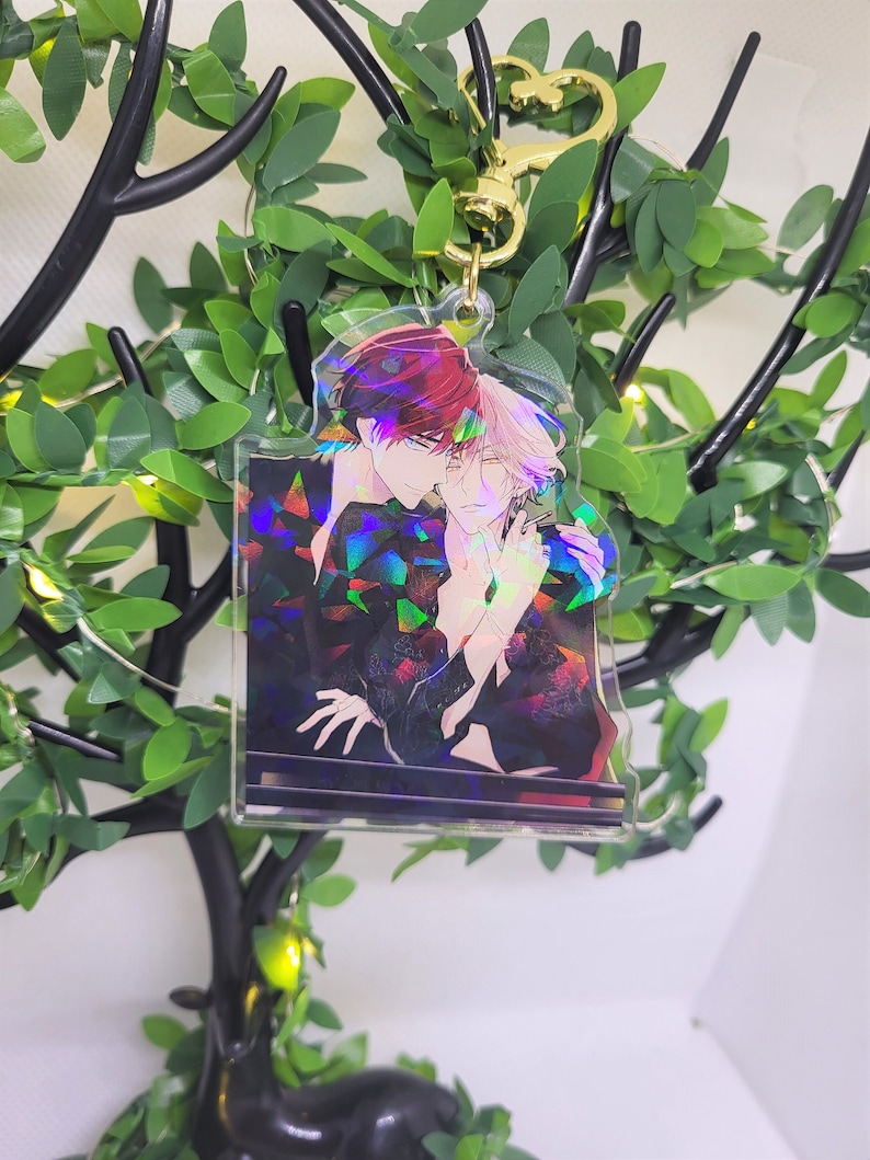 Op de afbeelding: Een heldere acryl sleutelhanger met een kleurrijke illustratie van twee anime-achtige personages die elkaar omarmen. De sleutelhanger heeft een goudkleurige hartvormige sluiting en een holografisch effect. De achtergrond bevat kunstgroen en een decoratieve boom.