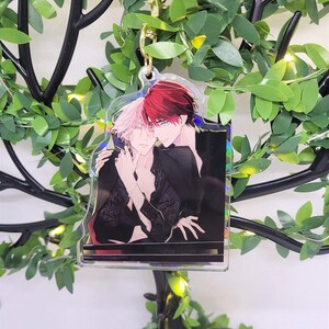 Op de afbeelding: Een heldere acryl bedel met een anime-stijl illustratie van twee figuren die elkaar omarmen. De bedel heeft een goudkleurige sluiting. De illustratie toont twee figuren met rood en roze haar, gekleed in zwarte kleding.