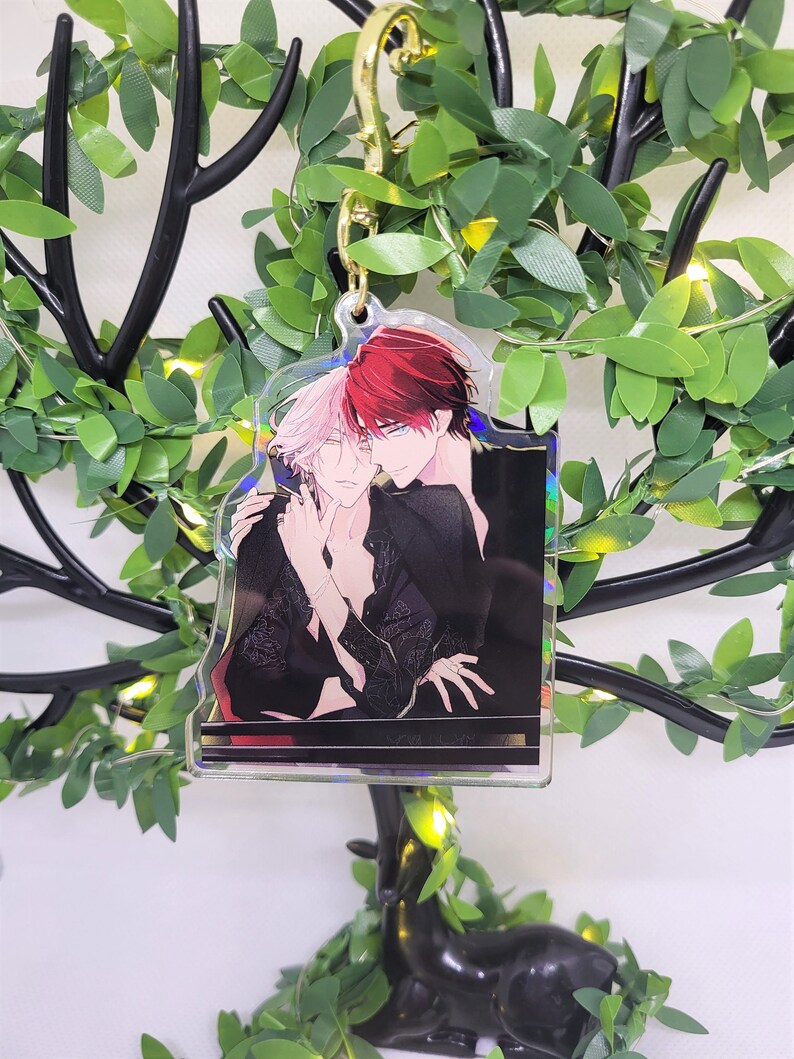Op de afbeelding: Een heldere acryl bedel met een anime-stijl illustratie van twee figuren die elkaar omarmen. De bedel heeft een goudkleurige sluiting. De illustratie toont twee figuren met rood en roze haar, gekleed in zwarte kleding.