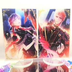 May include: Two acrylic standees with anime-style illustrations of a man and a woman. The woman has pink hair and wears a black dress. The man has blond hair and wears a dark uniform with gold accents. Both standees are on clear bases.