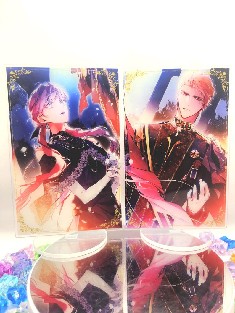 May include: Two acrylic standees with anime-style illustrations of a man and a woman. The woman has pink hair and wears a black dress. The man has blond hair and wears a dark uniform with gold accents. Both standees are on clear bases.