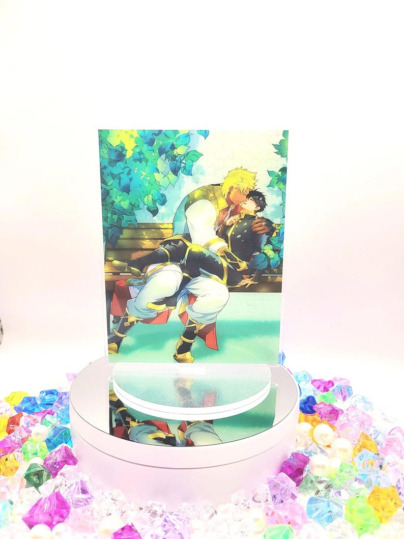 May include: A colorful illustration of two men embracing on a bench under a leafy tree. The man in the front is wearing a black and gold outfit, while the man in the back is wearing a white and gold outfit. The illustration is printed on a white card.