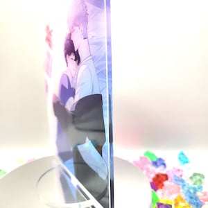 May include: A clear acrylic print featuring a soft illustration of two figures embracing. The artwork has a pastel color palette with blue, purple, and white hues. The print is displayed on a white rotating stand, with colorful decorative stones in the background.