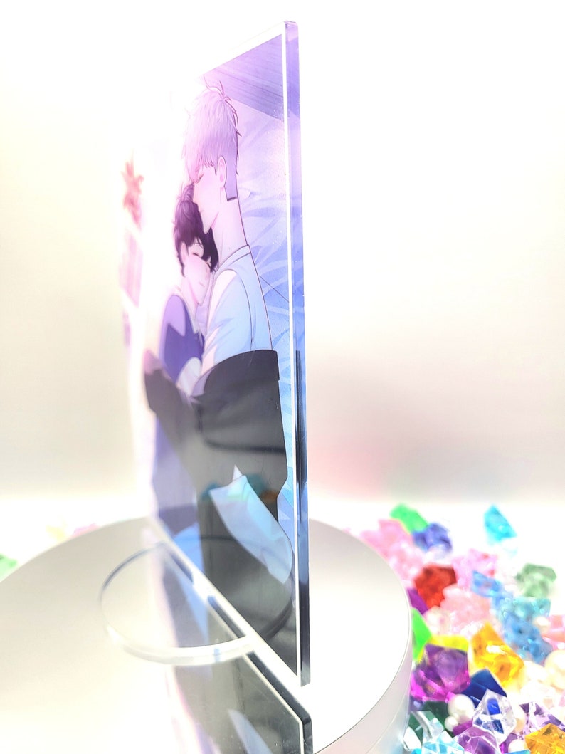 May include: A clear acrylic print featuring a soft illustration of two figures embracing. The artwork has a pastel color palette with blue, purple, and white hues. The print is displayed on a white rotating stand, with colorful decorative stones in the background.