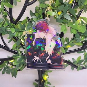 Op de afbeelding: Een heldere acryl bedel met een anime-stijl illustratie van twee mannen die elkaar omarmen. De bedel heeft een goudkleurige sluiting en een holografisch effect op de kleding. De achtergrond bevat een decoratieve boom met groene bladeren.