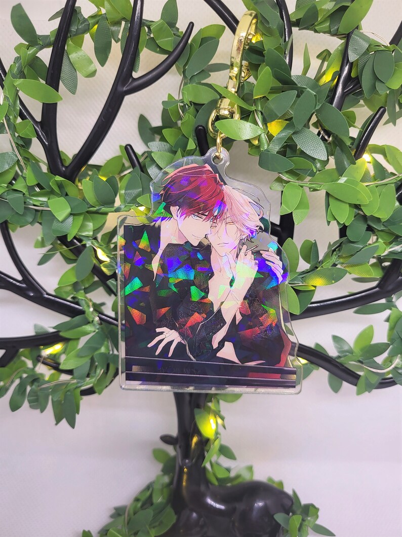 Op de afbeelding: Een heldere acryl bedel met een anime-stijl illustratie van twee mannen die elkaar omarmen. De bedel heeft een goudkleurige sluiting en een holografisch effect op de kleding. De achtergrond bevat een decoratieve boom met groene bladeren.