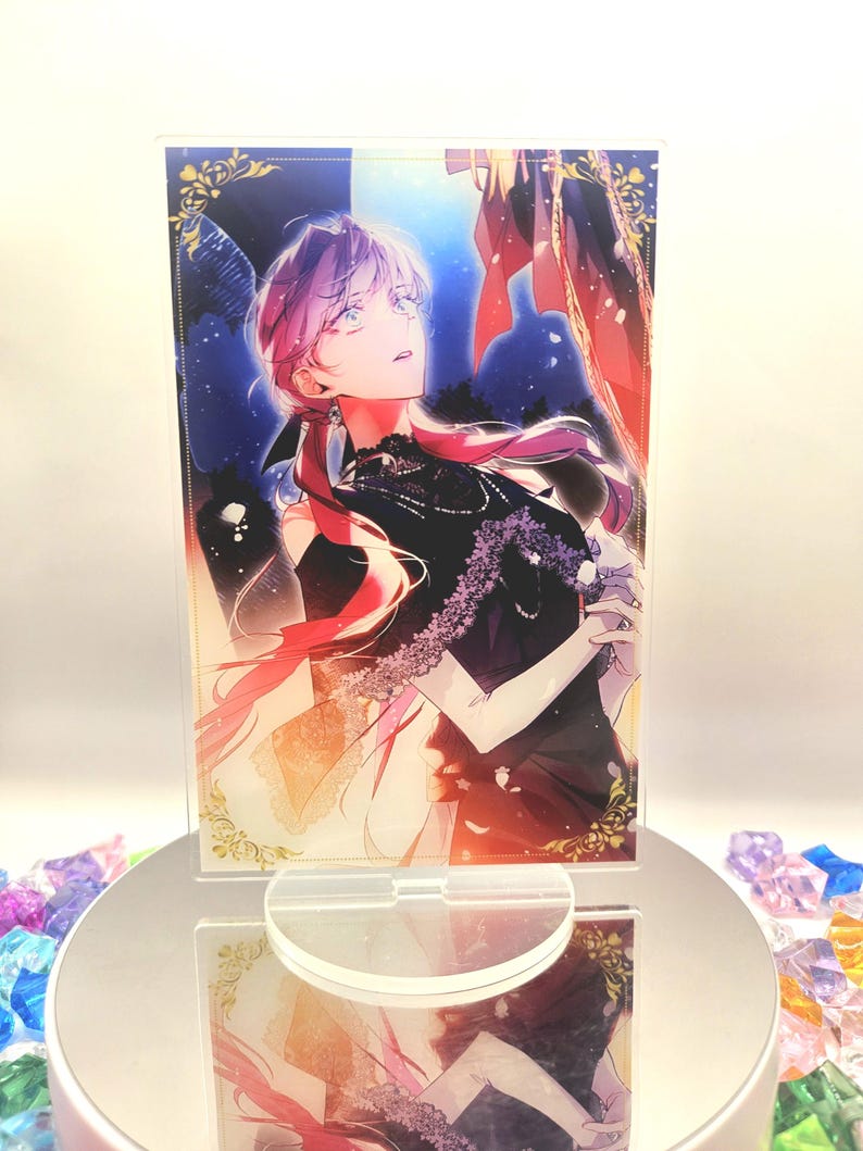 May include: A clear acrylic stand with a vibrant anime-style illustration of a person in a black dress and pink hair. The artwork has a dramatic, fantasy theme, set on a reflective base.