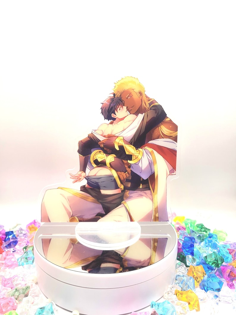 May include: A white acrylic stand with a clear circular platform. The platform features a printed image of two men embracing. The man in the front is wearing a gold and white outfit, while the man in the back is wearing a black and blue outfit.
