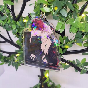 Op de afbeelding: Een heldere acryl sleutelhanger met een goudkleurige sluiting, met een anime-stijl illustratie van twee personages die elkaar omarmen. De sleutelhanger heeft een holografisch effect. De achtergrond is een decoratief arrangement van groene bladeren en lichtjes.