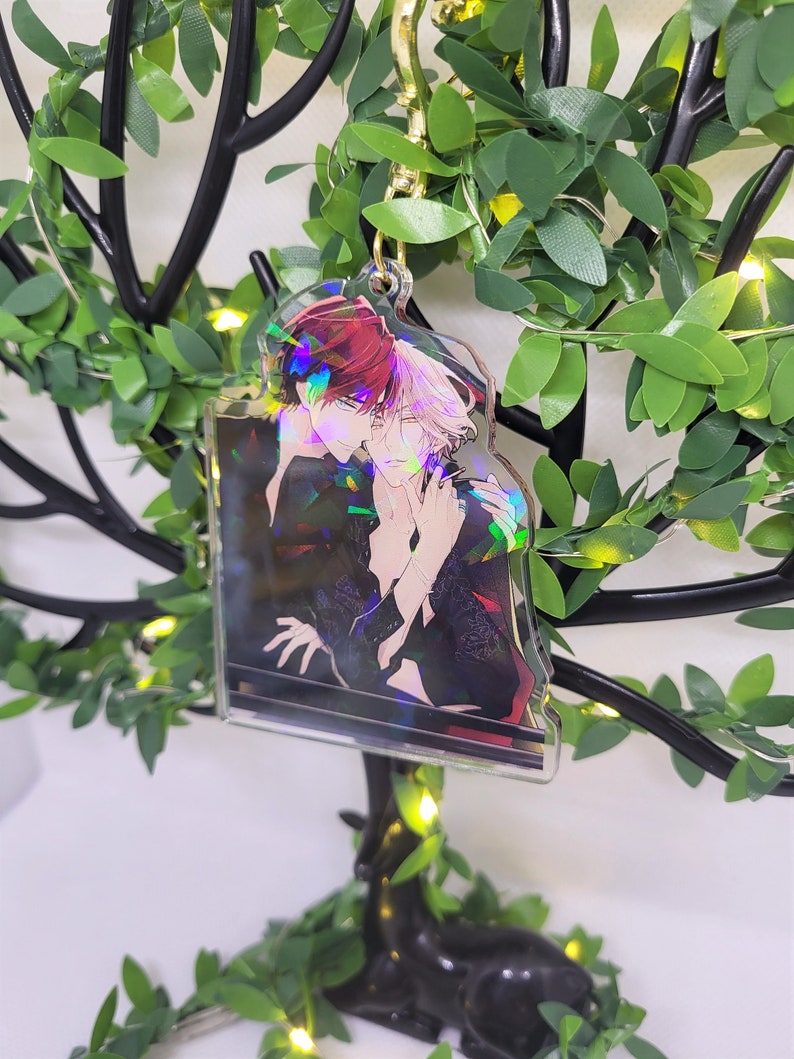 Op de afbeelding: Een heldere acryl sleutelhanger met een goudkleurige sluiting, met een anime-stijl illustratie van twee personages die elkaar omarmen. De sleutelhanger heeft een holografisch effect. De achtergrond is een decoratief arrangement van groene bladeren en lichtjes.