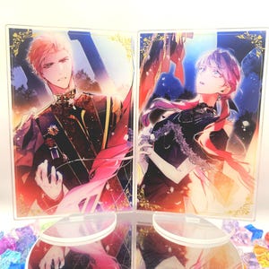 May include: Two acrylic display stands with anime-style illustrations of a man and a woman. The man is in a dark uniform with gold details, the woman in a black dress. Both have colourful hair and are set against a vibrant background.