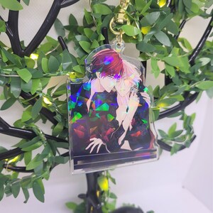 Op de afbeelding: Een heldere acryl sleutelhanger met een holografische afbeelding van twee anime-stijl figuren die elkaar omarmen. De sleutelhanger heeft een goudkleurige sluiting en wordt gepresenteerd tegen een achtergrond van kunstgroen en een decoratieve boom.