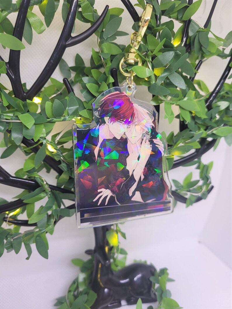 Op de afbeelding: Een heldere acryl sleutelhanger met een holografische afbeelding van twee anime-stijl figuren die elkaar omarmen. De sleutelhanger heeft een goudkleurige sluiting en wordt gepresenteerd tegen een achtergrond van kunstgroen en een decoratieve boom.