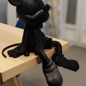 Può includere: Una statuetta nera di Topolino seduta sul bordo di un tavolo di legno chiaro. La statuetta ha grandi orecchie, un viso rotondo e indossa pantaloncini e scarpe. Le gambe della statuetta penzolano dal bordo del tavolo.
