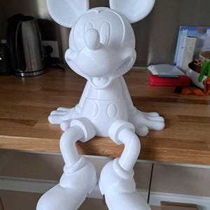 Peut inclure: Une figurine blanche de Mickey Mouse, avec de grandes oreilles rondes et un visage souriant. La figurine est assise, les jambes pendantes sur le bord d'une surface en bois. La figurine est faite d'un matériau lisse et solide, d'environ 30 cm de haut.