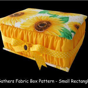 May include: A yellow fabric box with a sunflower print lid and a yellow ribbon with white polka dots. The box has a gathered fabric edge and is a small rectangle.