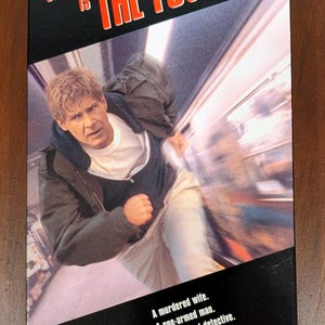 May include: VHS tape for the movie "The Fugitive" starring Harrison Ford. The cover features a photo of Ford running, with the title in red and white lettering. Text on the bottom reads: "A murdered wife. A one-armed man. An obsessed detective. The chase begins."