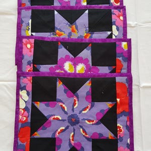 May include: Four purple and black quilted placemats with a floral pattern. The placemats are made of fabric and have a star design in the centre.