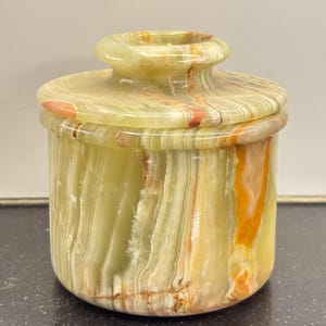 May include: A cylindrical jar with a lid, crafted from green onyx. The jar features bands of color, including shades of yellow, orange, and brown. The lid has a rounded top with a small, circular handle. The jar is likely used for storage or display.