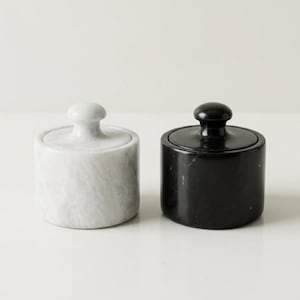 May include: Two cylindrical containers with matching lids, one crafted from white marble with grey veining and the other from solid black stone. Each container features a smooth finish and a small, rounded knob handle on top of the lid.