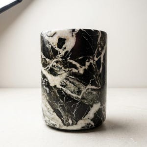 May include: A cylindrical vessel crafted from polished stone, featuring a deep black base color with prominent, swirling white and dark green veining throughout its surface. The object has a smooth, rounded top edge and a solid, upright form.
