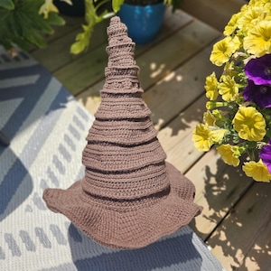 Peut inclure: Un chapeau marron en crochet avec un large bord et une couronne conique haute et étagée. Le chapeau est fait d'un fil doux et texturé. Le chapeau est posé sur une surface à motifs.